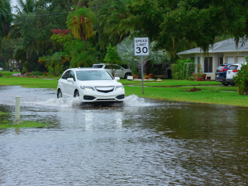 Heavy flooding and storm surge in residential neighborhood with a car driving through deep splashing water in the flooded street in front of houses with Speed Limit sign on side of the road.