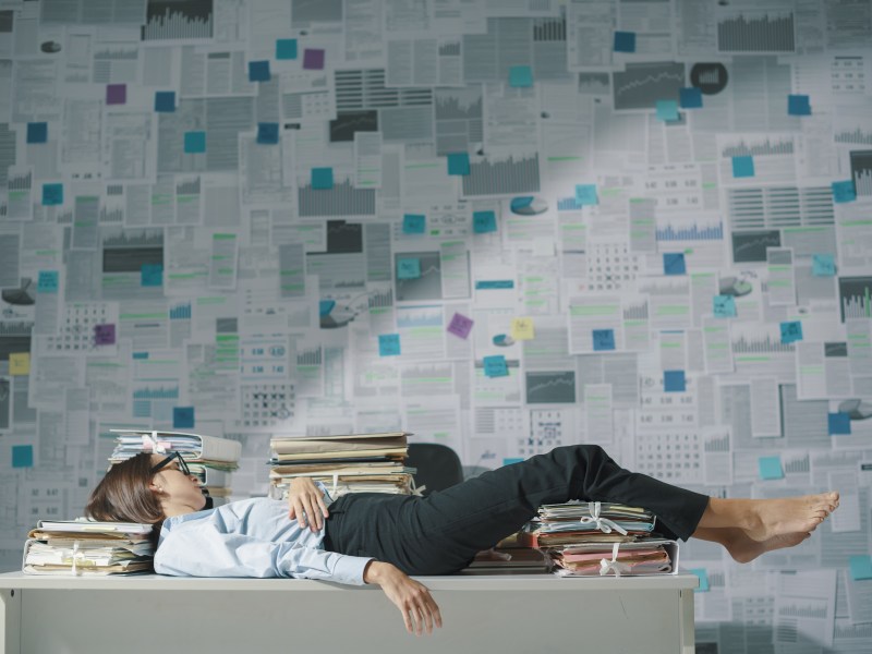 Exhausted business executive lying down on the desk and sleeping, wall covered with financial reports in the background, overtime work and deadlines concept