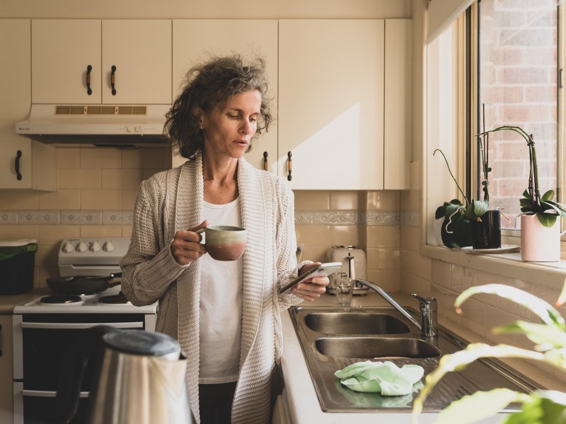 Mature woman looking at phone and holding up in kitchen with matte filter effect