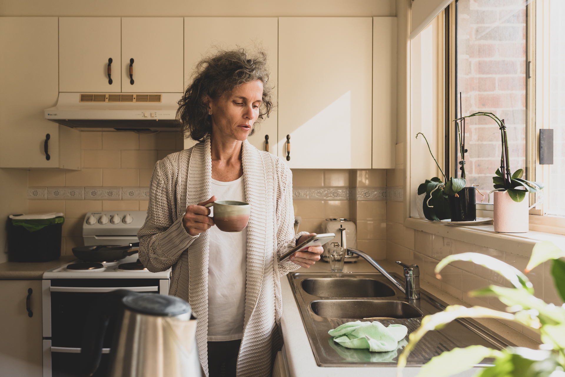 Mature woman looking at phone and holding up in kitchen with matte filter effect