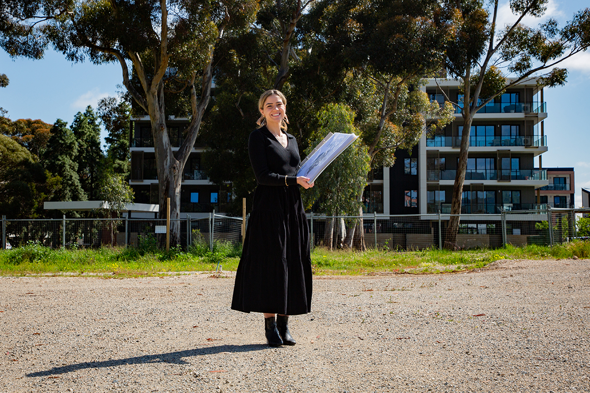 woman in long black dress smiling, standing in front of apartment buildings with large gum tree in the garden