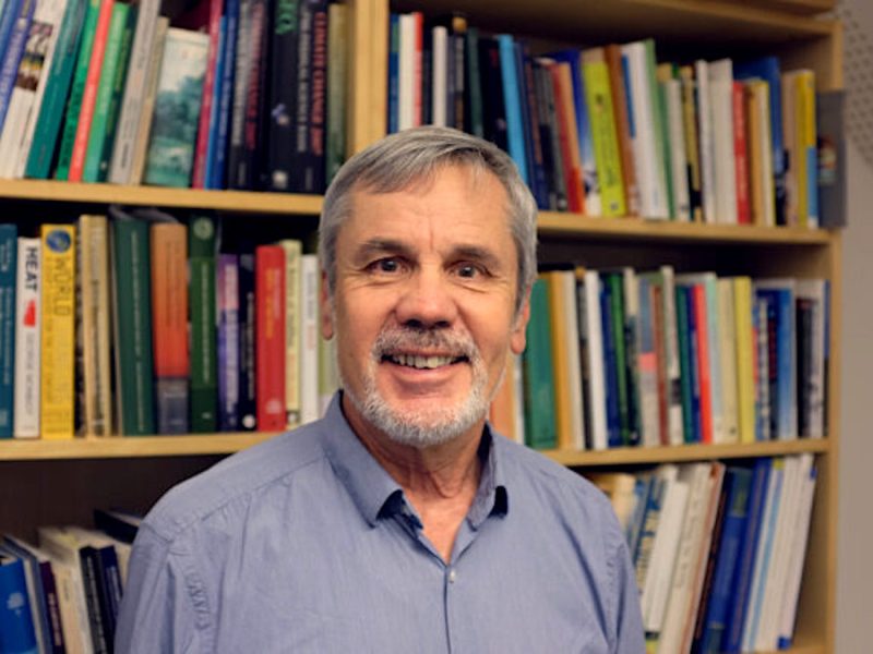 Older caucasian man smiling at the camera, wearing a blue shirt, with a bookcase full of books on the wall behind him.