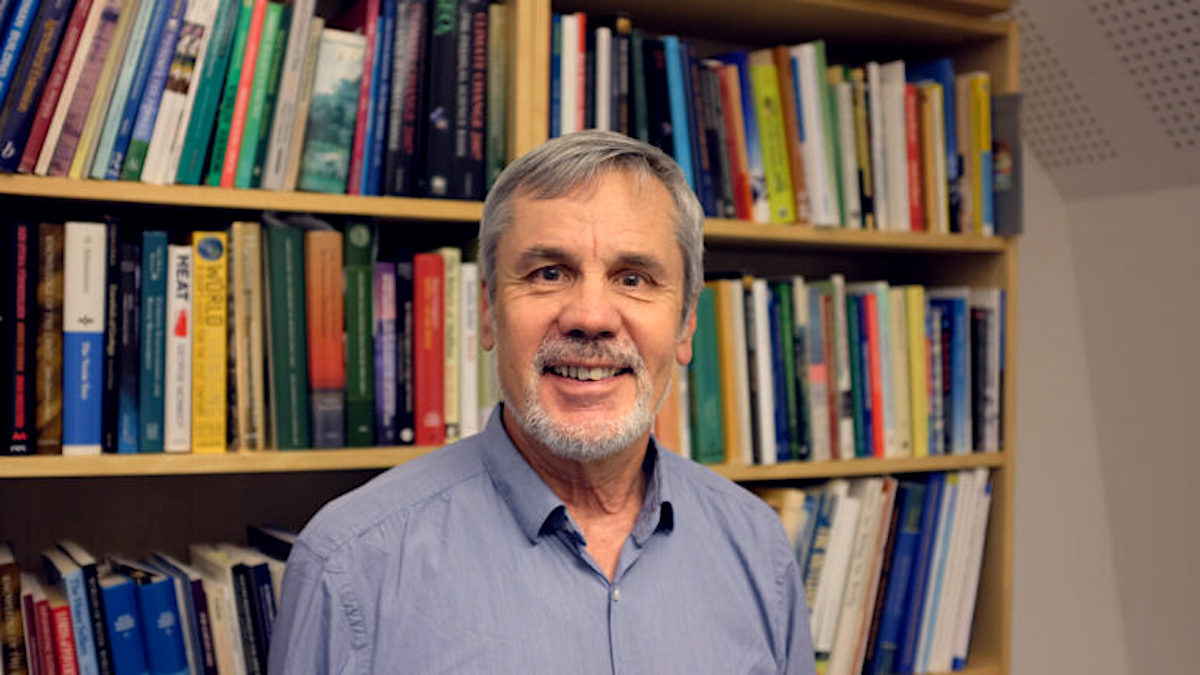 Older caucasian man smiling at the camera, wearing a blue shirt, with a bookcase full of books on the wall behind him.