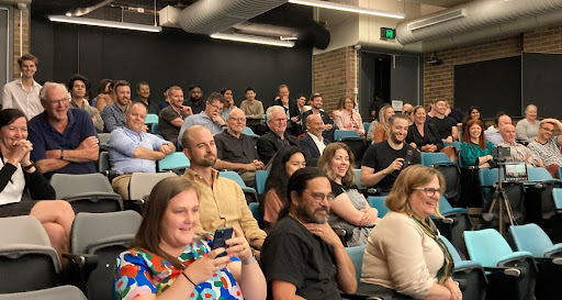 Photo of audience sitting in a theatre watching an unseen event on stage. People are smiling and clapping and laughing 