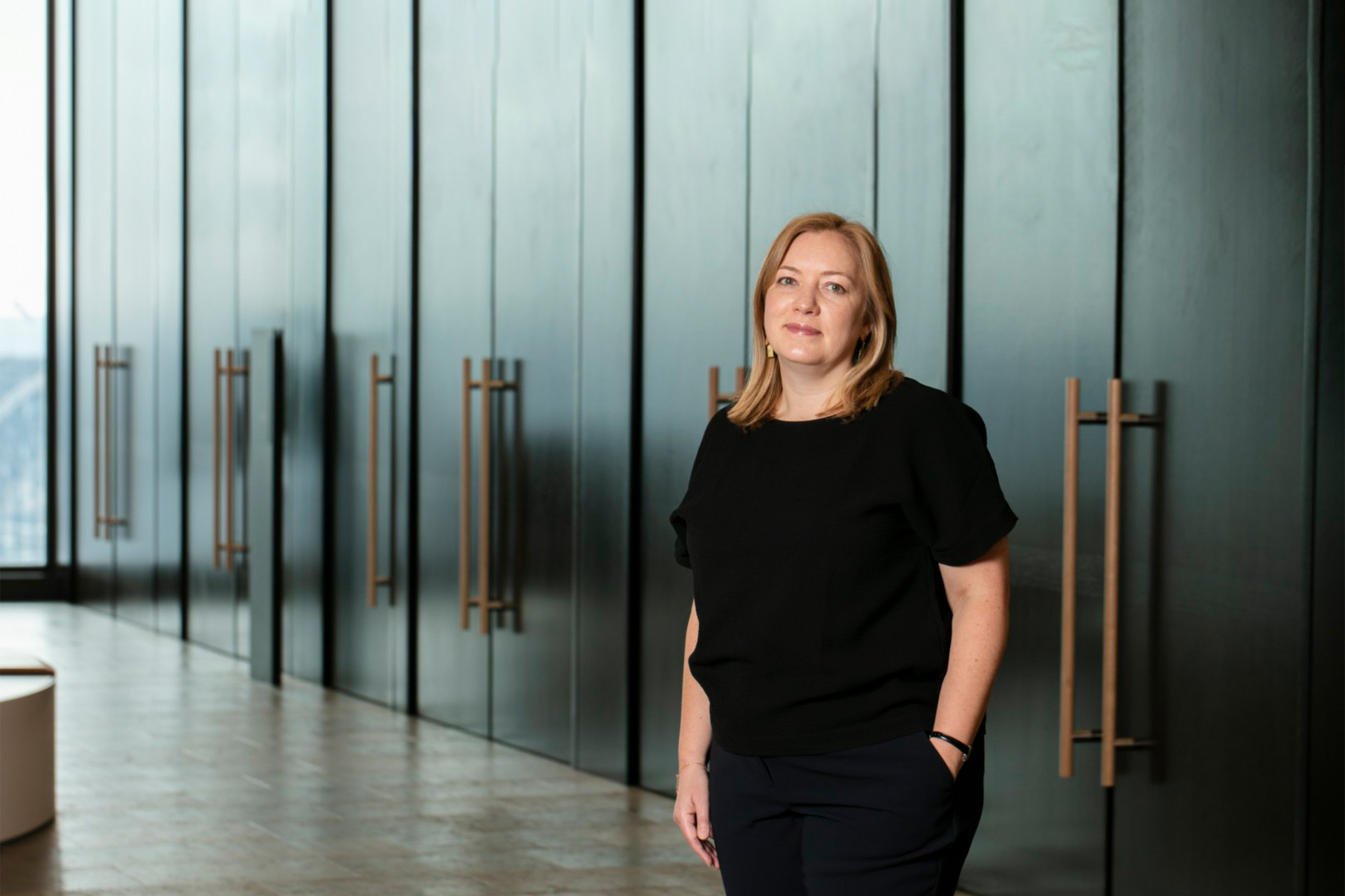 Professional woman with medium length blonde hair in black dress, standing against a dark corporate backdrop with hands in pockets and a serious expression
