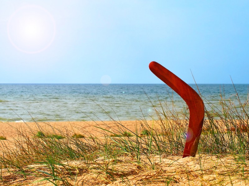 Landscape with boomerang on overgrown sandy beach against blue sea and sky.