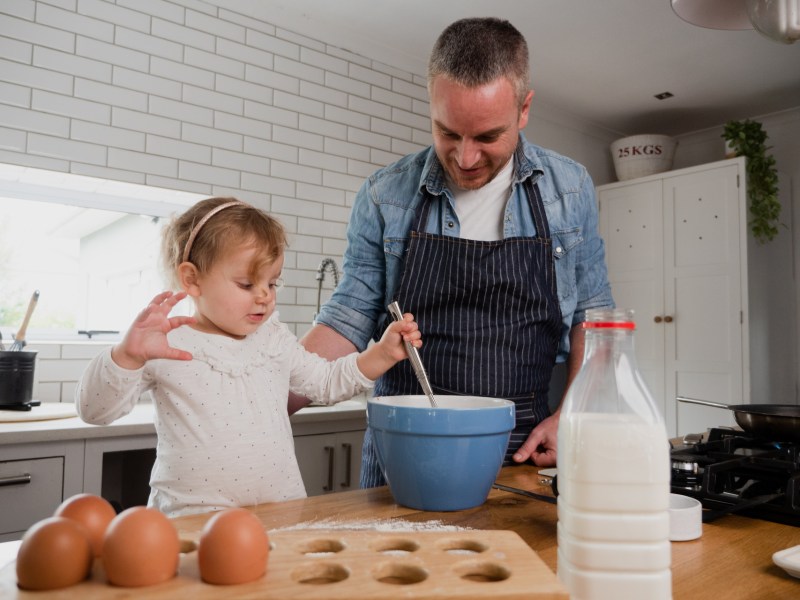 Caucasian father and toddler daughter happily mixing ingredients in bowl together, in kitchen at home using a gas stove