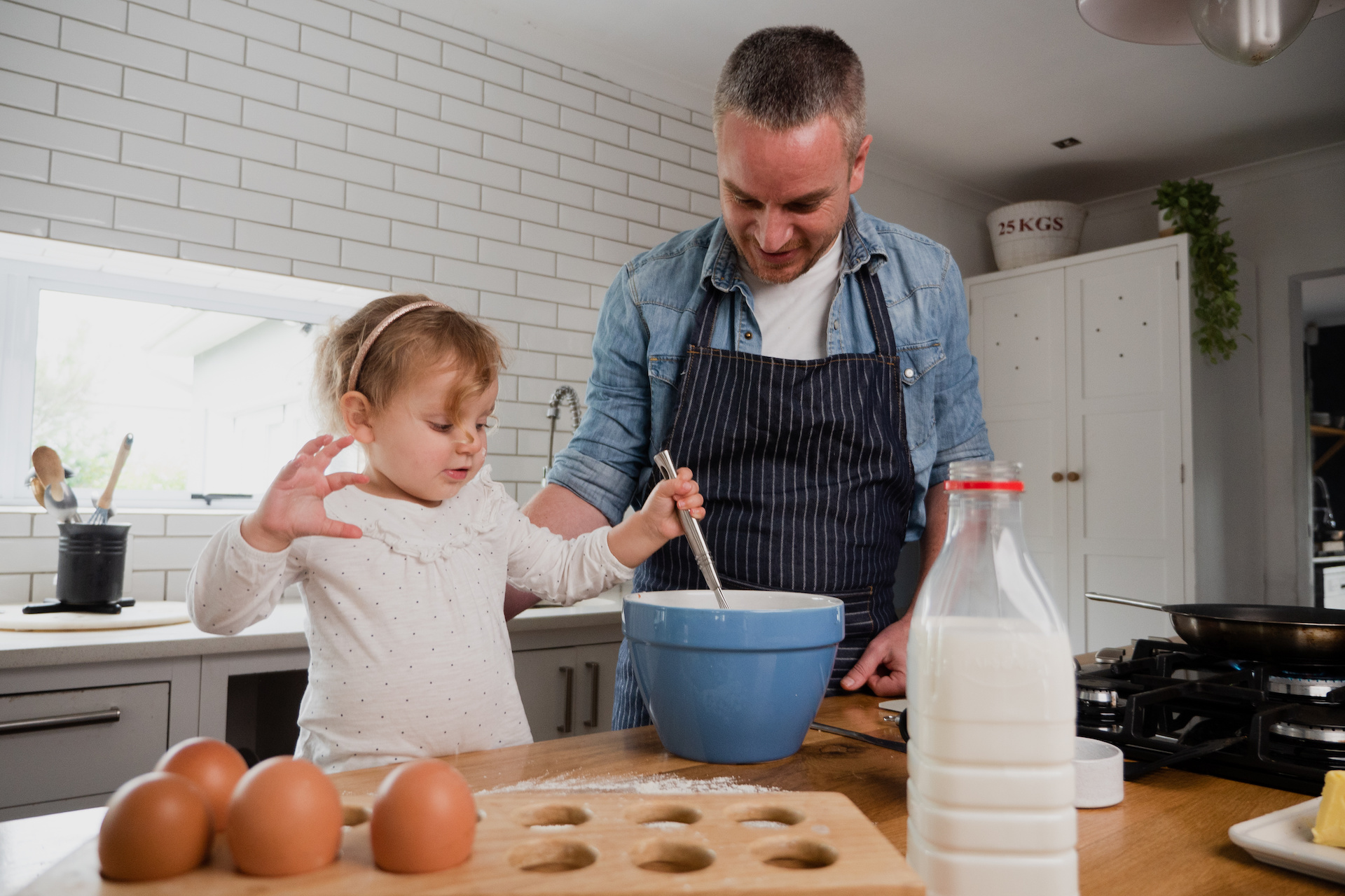 Caucasian father and toddler daughter happily mixing ingredients in bowl together, in kitchen at home using a gas stove