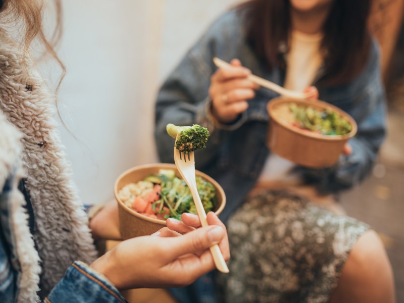 Two millennial women female friends sitting outdoors eating takeaway food, laughing and having fun. Food delivery and takeout.