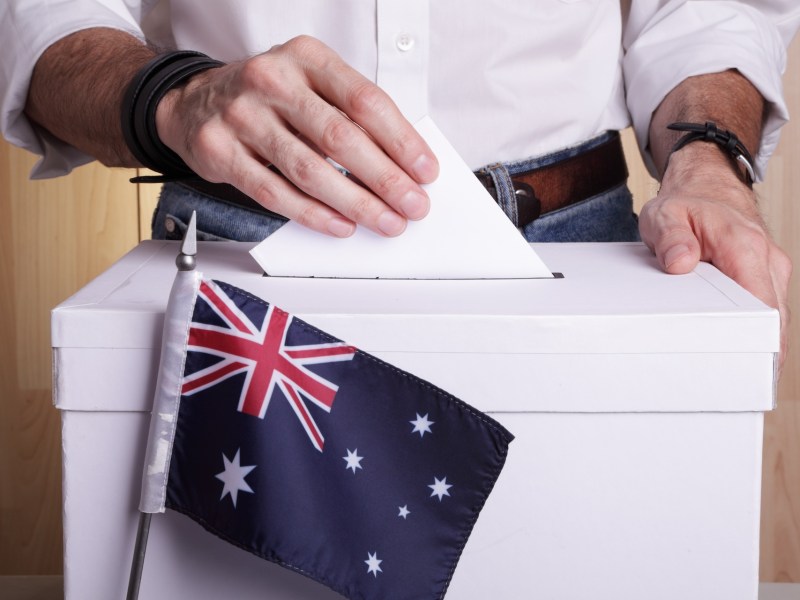 An Australian citizen inserting a ballot into a ballot box. Australia flag in front of it